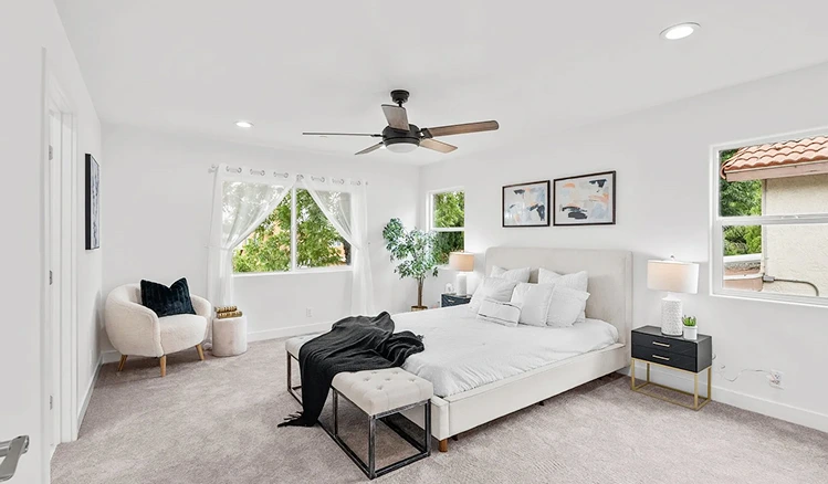 A modern bedroom with a white bed, plush white pillows, and a black throw blanket. The room is decorated with abstract art, a ceiling fan, and a large window overlooking a lush green garden.